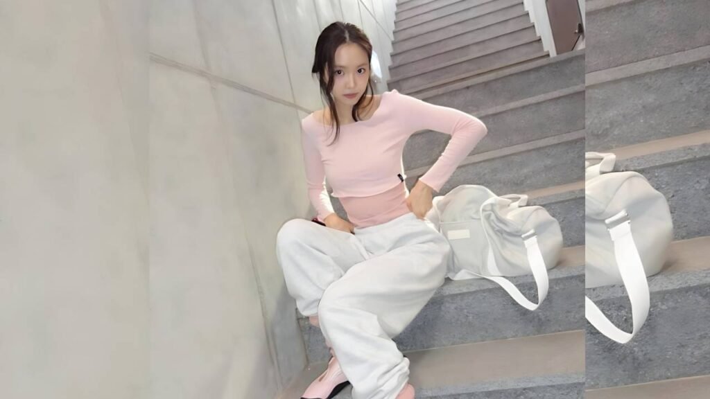 Son Na-eun in pale pink top and white baggy pants sits on gray stairs, with a light gray duffel bag beside her.