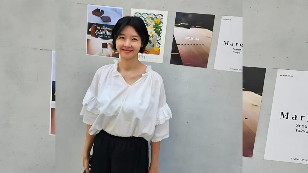 Song Seon-mi with short black hair in a white ruffled blouse and black skirt stands against a gray wall with fashion posters