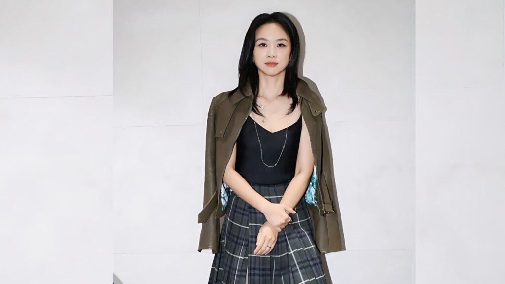 Tang Wei with shoulder-length black hair stands against pale tiled wall, wearing olive-green jacket over a black top and plaid skirt, hands clasped.