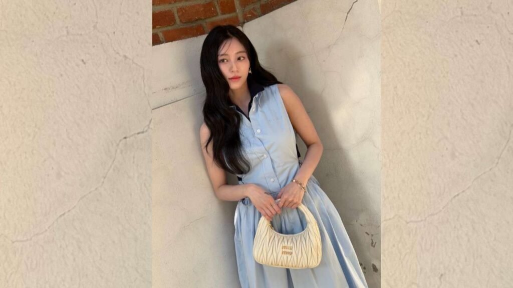 Kang Min-ah in a sleeveless light blue dress leans against a pale textured wall, holding a cream handbag and smiling softly.