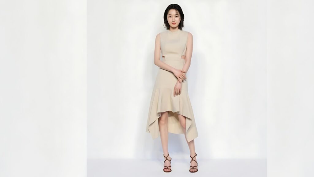 Won Ji-an in a beige sleeveless dress with a high-low ruffled hem poses against a white studio backdrop, hands folded, strappy sandals.