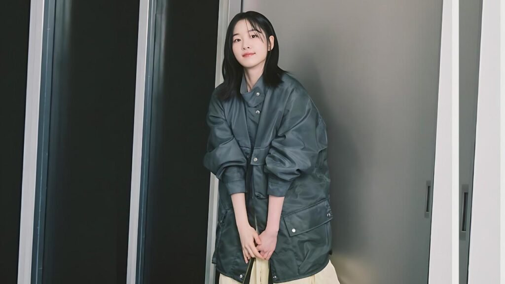 Won ji an (지안) with shoulder-length black hair in oversized dark green jacket, hands folded, posing against grey paneled wall.