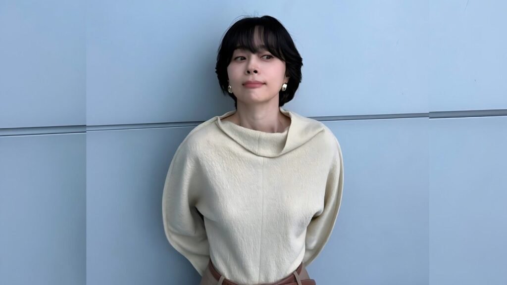Woo Hee-jin with short black hair in a cream oversized sweater stands against a pale blue wall, looking softly to the side.