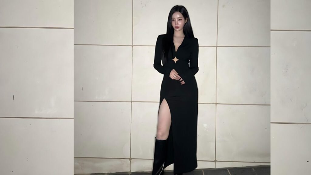 Yeonwoo in a black, long-slit dress with a star cut-out, knee-high boots, posing against pale tiled wall.
