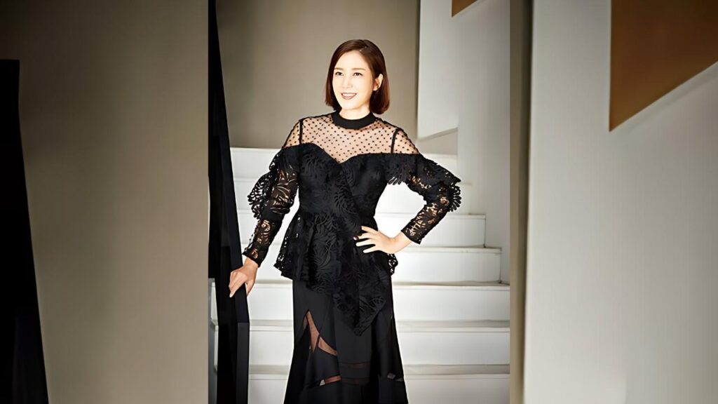 Yoo Ho-jeong in a black lace dress stands on a bright white staircase, one hand on her hip, the other on the railing.