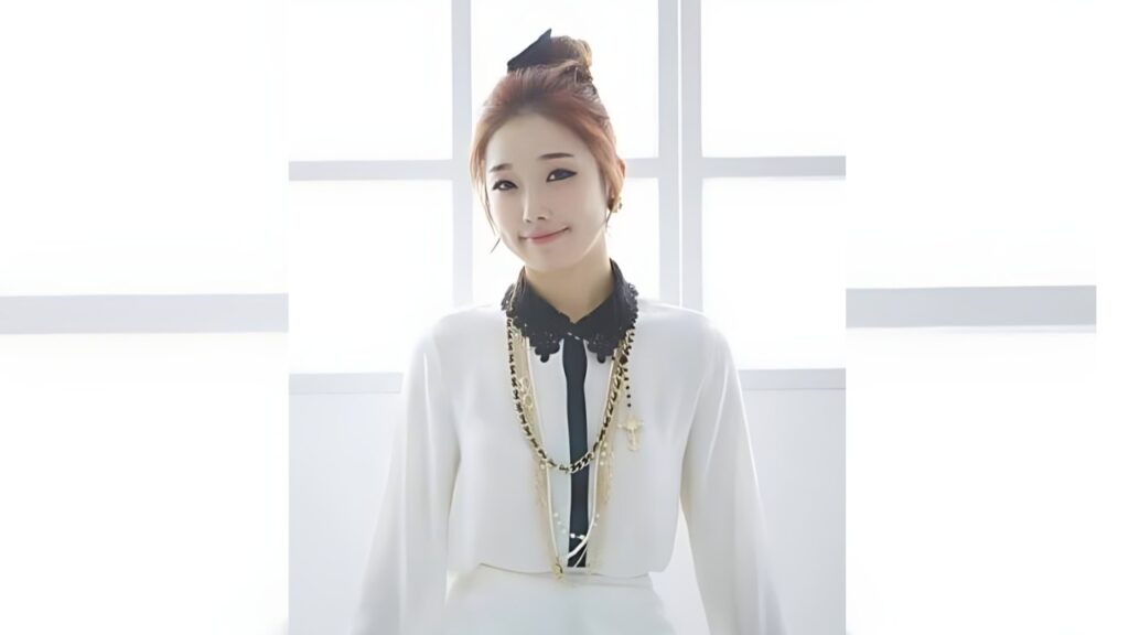 Yoo Pool-ip (유풀잎) in a white blouse with a black lace collar, black tie, and gold chains, posed before a bright window backdrop.