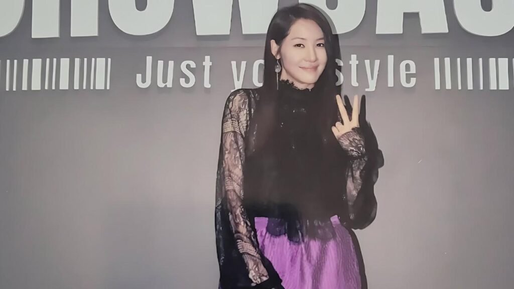 Yoon Ji-min with long dark hair in a sheer black lace top and purple skirt poses a peace sign against a gray wall.
