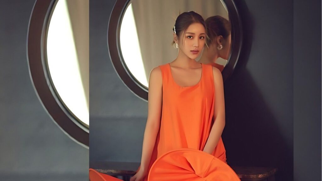 Yoon Jin-yi in a bright orange sleeveless dress sits against a dark wall beside a circular window; her reflection appears in the mirror behind her.