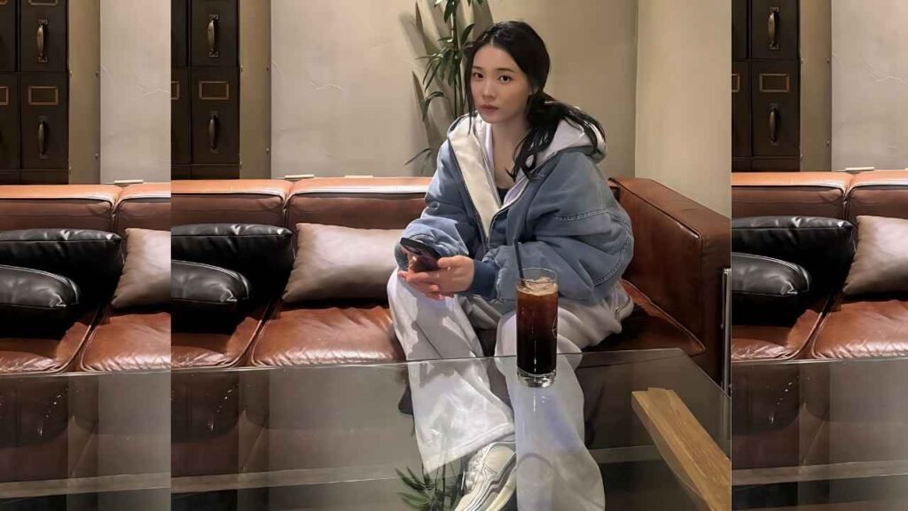 Yoon So-hee with a white hoodie, young woman sits on a brown leather couch, holding a smartphone; iced coffee on a glass table nearby.