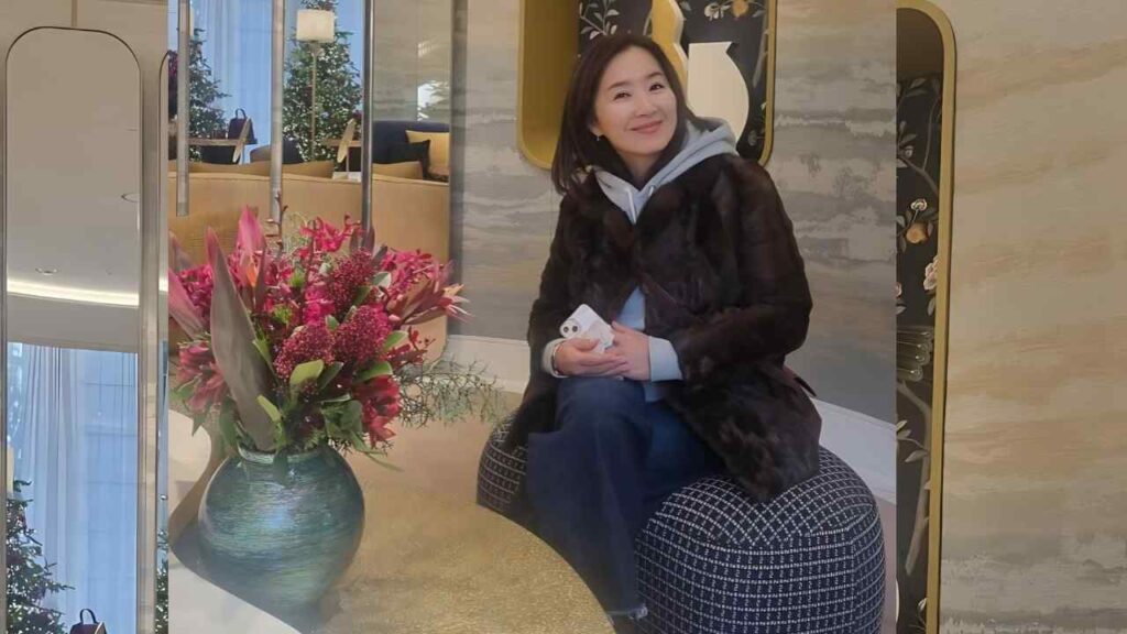 Yoon Yoo-sun in a dark fur coat and hoodie sits on a black-and-white patterned pouf, holding a phone, in a stylish lobby.