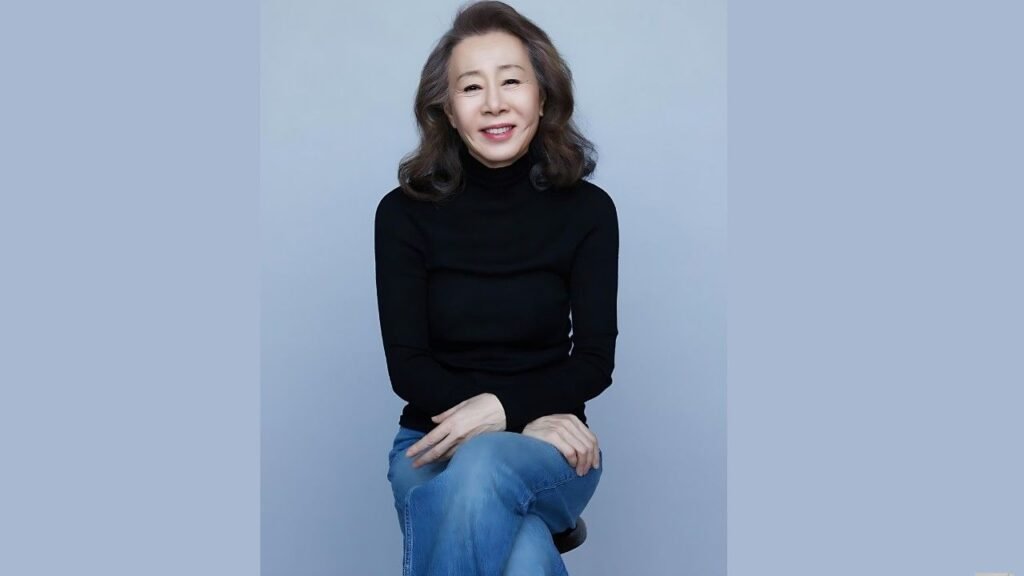 Youn Yuh-jung with shoulder-length hair sits in a black turtleneck and jeans against a pale blue backdrop.