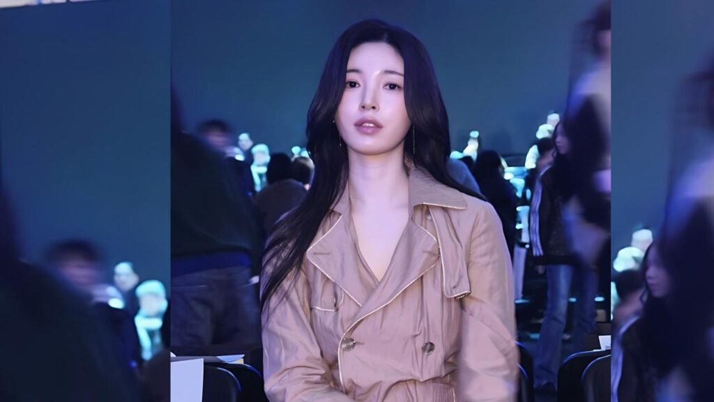 Yu Seung-ok (유승옥) with long dark hair sits center in a beige trench coat, gazing at the camera, blue-lit crowd blurred behind.