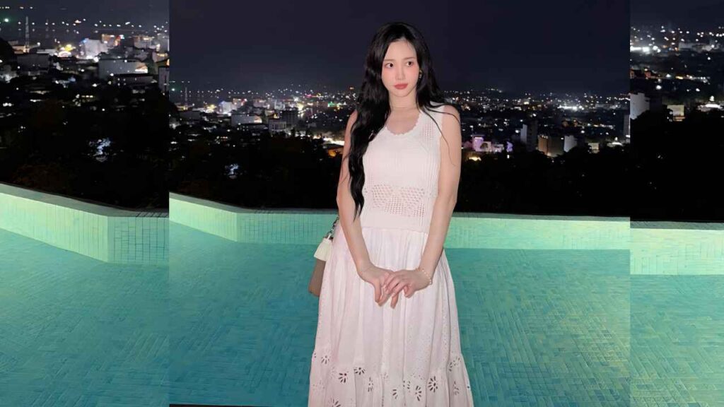 Yura with long dark hair in a white dress beside a turquoise pool, city lights behind, hands clasped.
