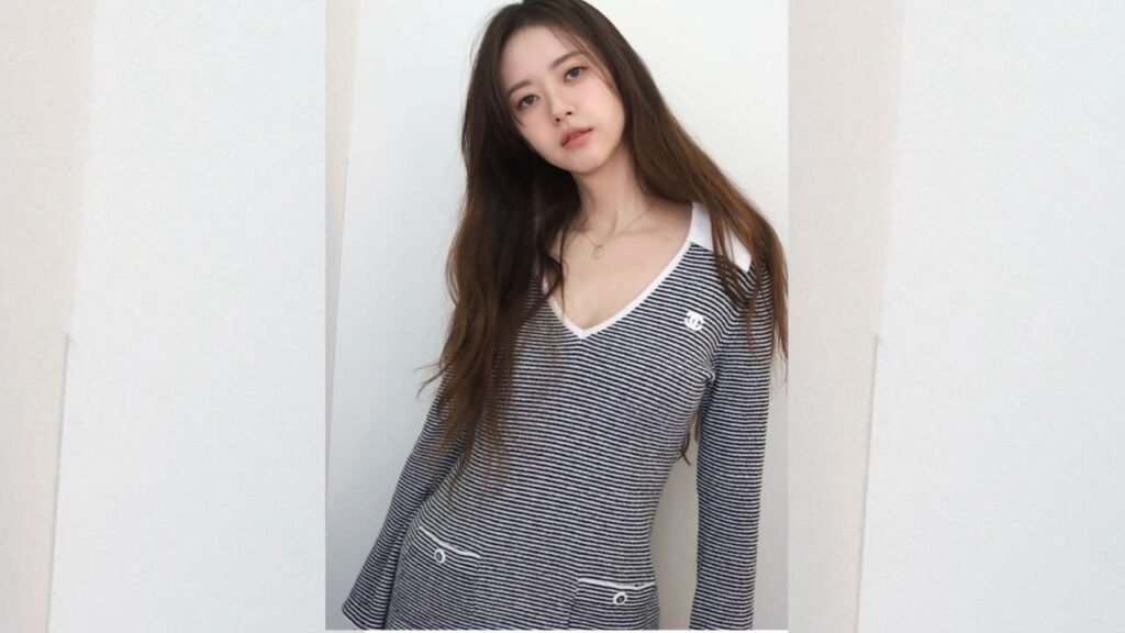 Go Ara with long, wavy hair wearing a black-and-white striped V-neck sweater with white trim, posing against pale walls.